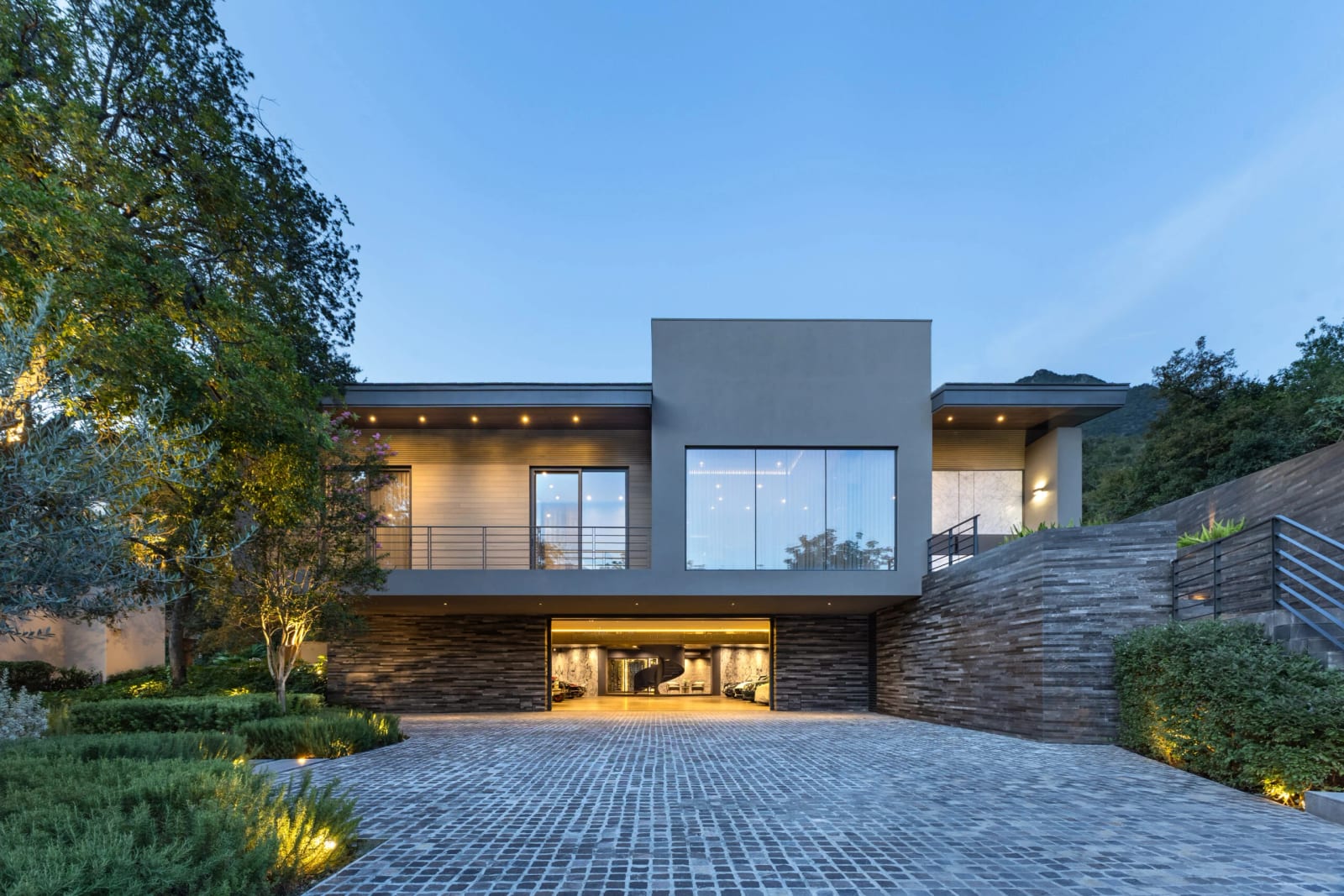 Fachada de Casa Saponaria, de GLR Arquitectos, al atardecer, con la cochera iluminada al centro y montañas al fondo