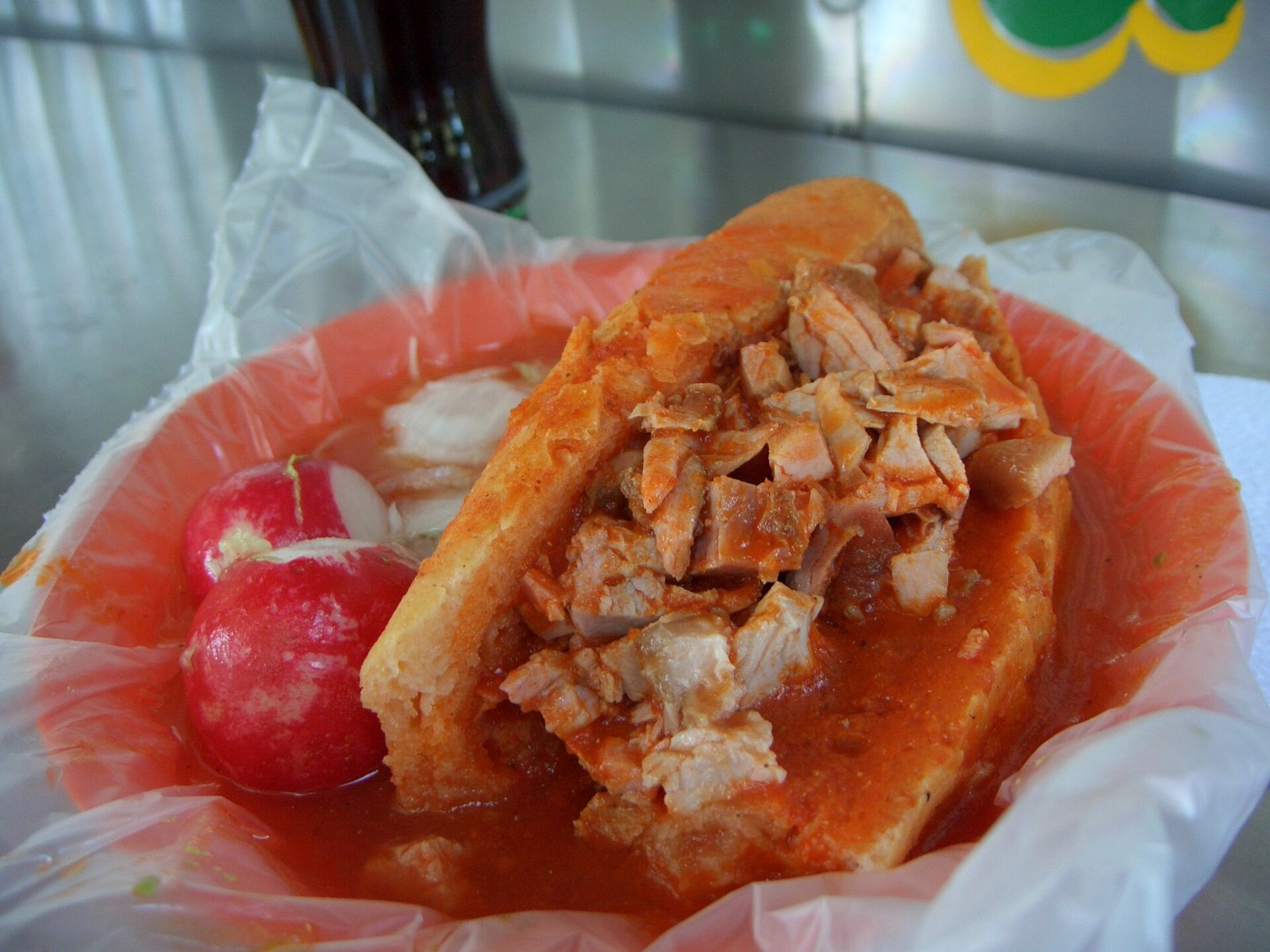 Torta ahogada de carnitas bañada en salsa roja, servida en un plato forrado con plástico junto a dos rábanos