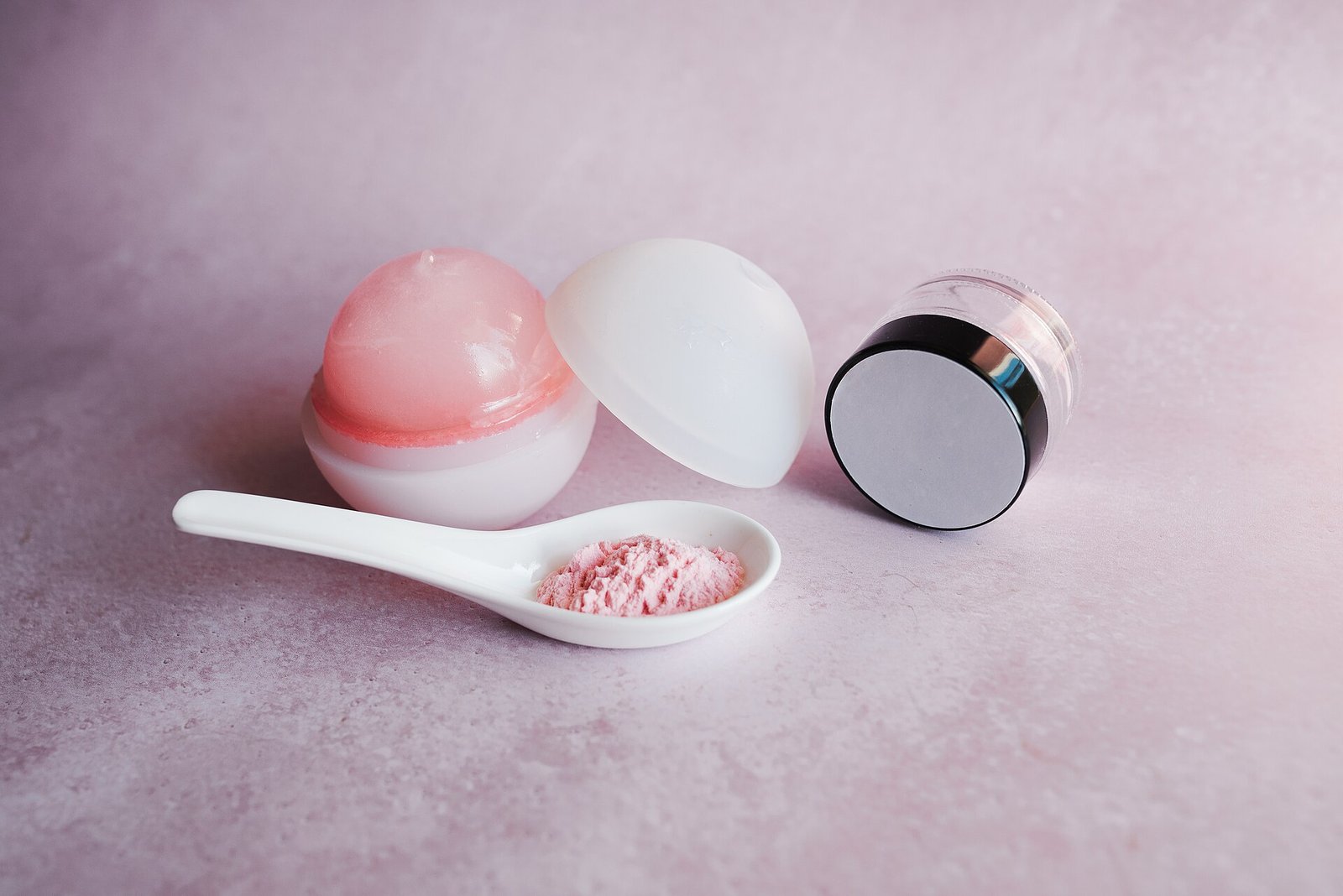 Frascos de productos para el cuidado de la piel y una cuchara con polvo rosa sobre un fondo rosa claro