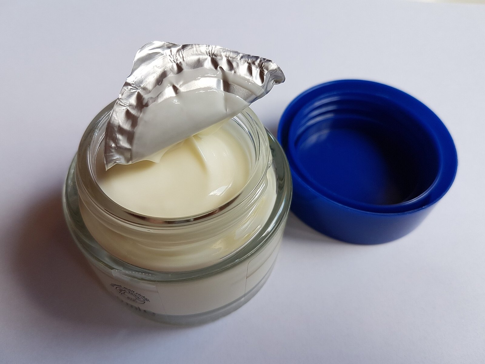 Frasco de vidrio abierto con crema cosmética blanca junto a su tapa azul